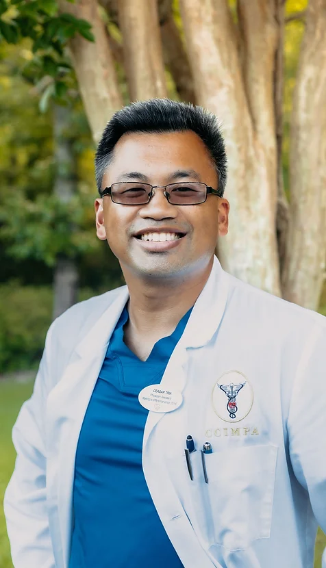Ceasar Tria, physicians assistant at Bloom Aesthetics and Wellness, poses outside smiling in a blue scrubs with a white lab coat over it.
