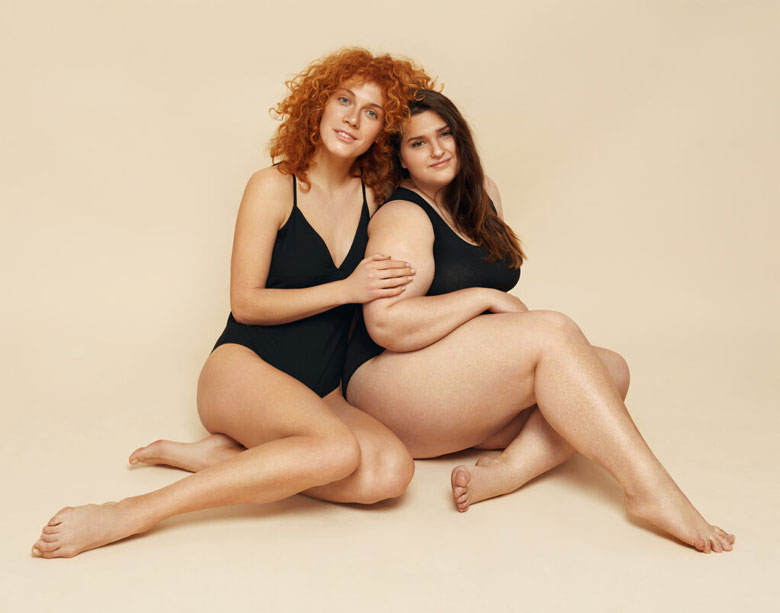 One beautiful woman with curly red hair sits next to a woman with long brown hair. One woman has her hand on the other's arm. Both women have natural bodies and wear black body suits to posing for the benefits of aesthetics treatment at Bloom Aesthetics and Wellness.