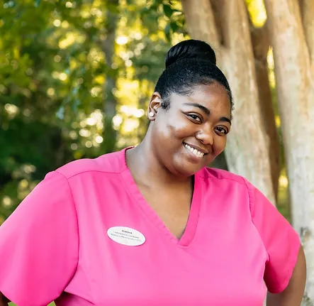 Porsha, Ceasar Tria's medical assistant at Bloom Aesthetics and Wellness poses outside smiling wearing pink scrubs.