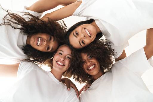 Four women of various ethnicities pose with heads together looking down at the camera with arms around each other's shoulders smile to model the testimonials section of the homepage for Bloom Aesthetics and Wellness.